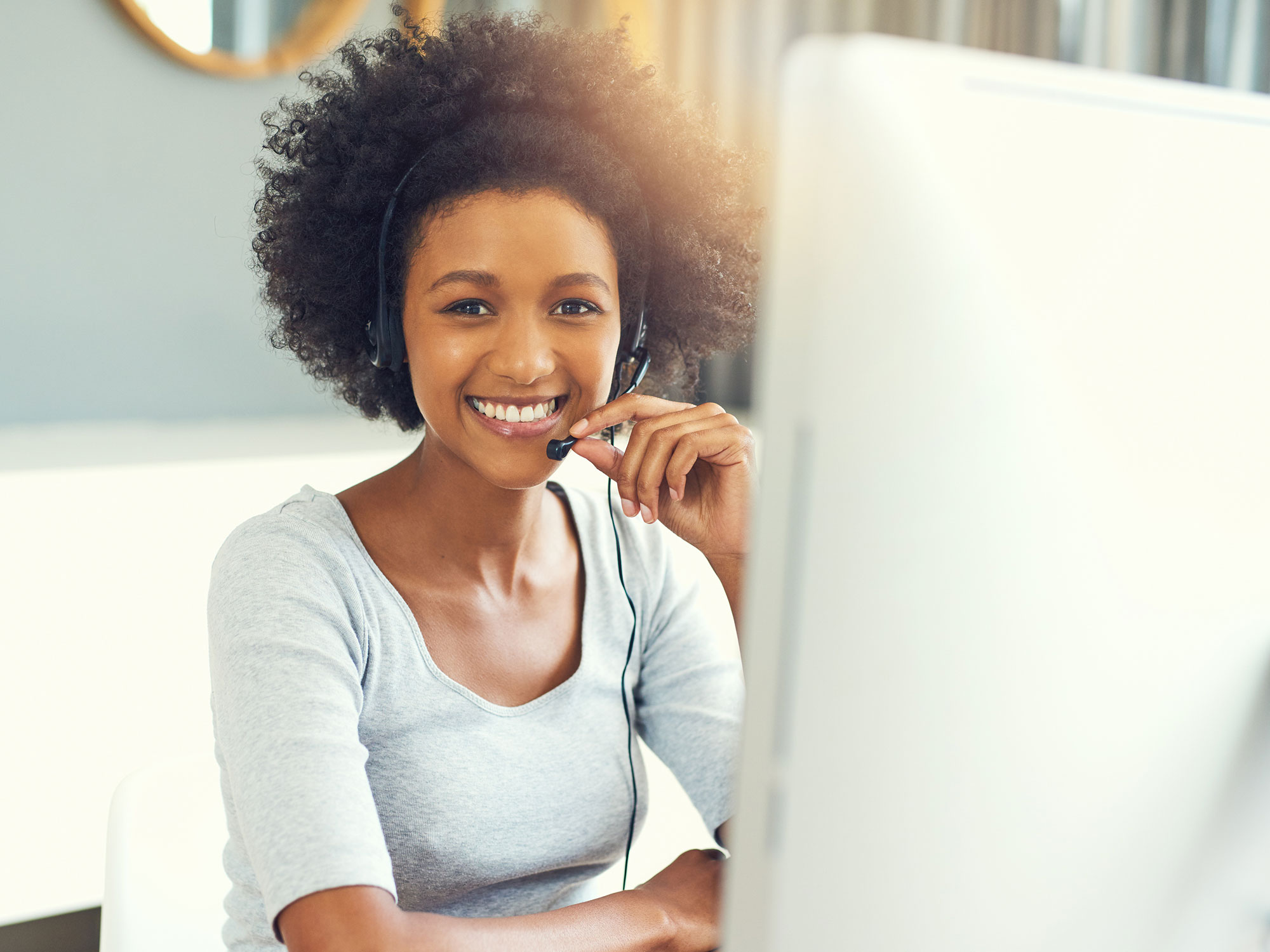 A young African American woman smiles at the camera, sitting at her computer and speaking on a headset for her work from home job